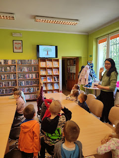 Sala biblioteczna. Pani bibliotekarka stoi z boku stolików przy których siedzą dzieci i pokazuje im prezentacje o drzewach wyświetlaną na ekranie telewizora.