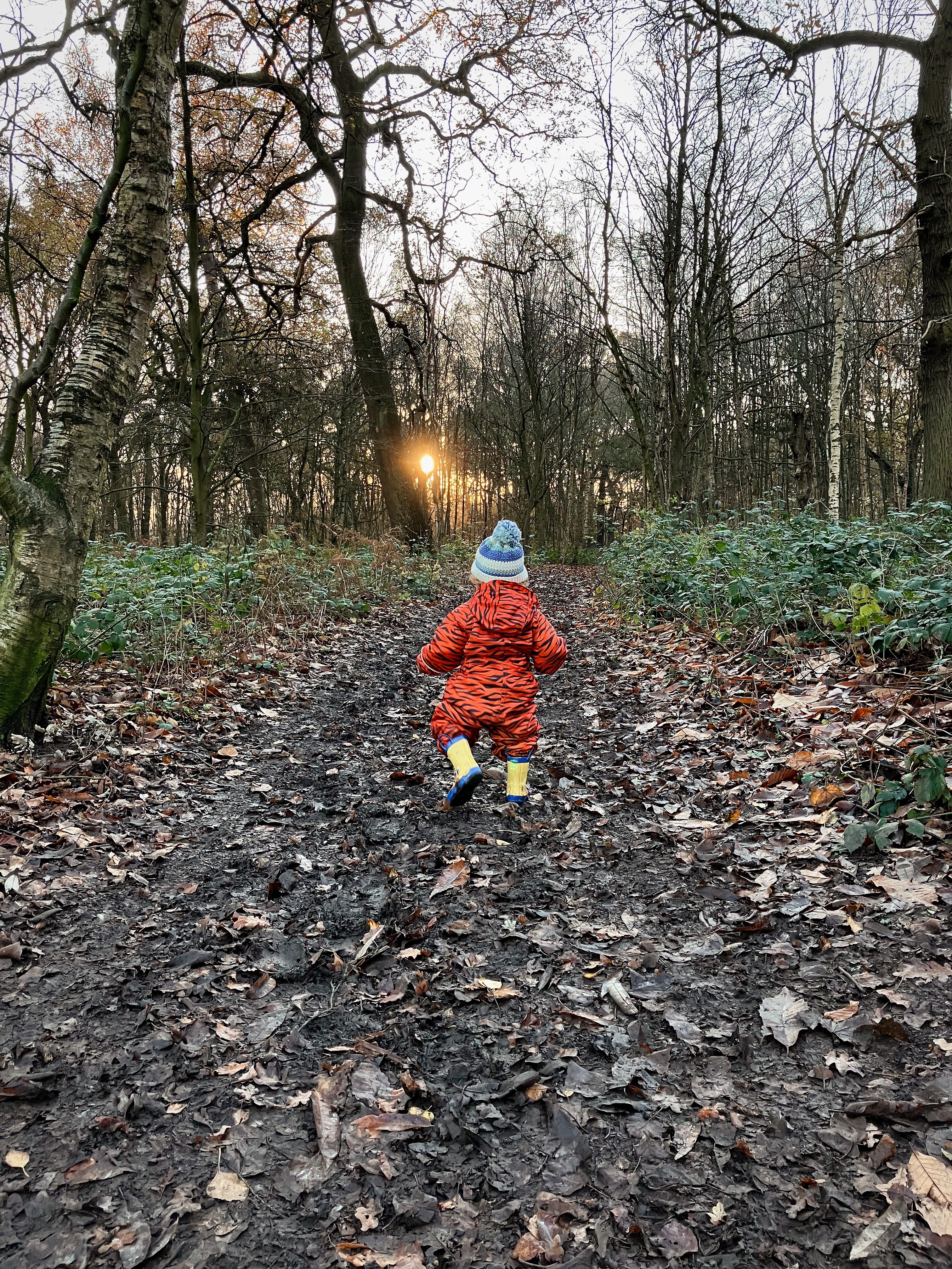 a toddler is running away into a forest wearing a tiger striped snowsuit and yellow wellies