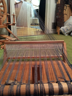 A small rigid heddle table loom, seen from just behind the cloth beam, with grey and a few teal warp threads stretching from the beam, through the heddle, and away into another room through a doorway, where they're tied to a pale wooden dowel suspended from a pillar on a white half-wall by an orange-and-turquoise strap. It's a long way.