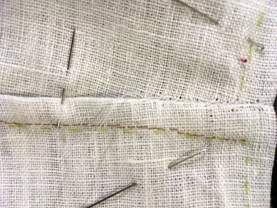 A close-up of a flat-felled seam in white linen, with a number of pins in very different directions, and bright green basting stitches running under and beside the tan stitching.