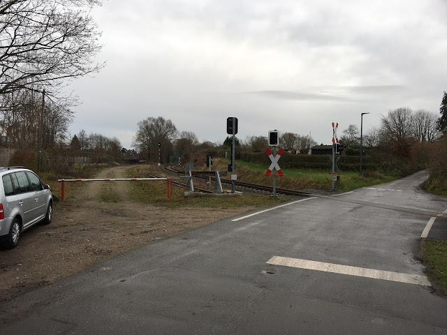 Hier geht der unbeschrankte Bahnübergang über das mit Beton oder Asphalt gefüllte Gleis und führt zwischen Schrebergärten weiter. Links steht ein silbergrauer Kombi, dann eine weiß-rote Schranke und ein Prellbock. Signale und Andreas-Kreuze sichern den Bahnübergang.