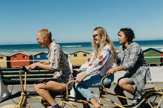 three women in a polyamorous triad on a bicycle built for 3, at a beach