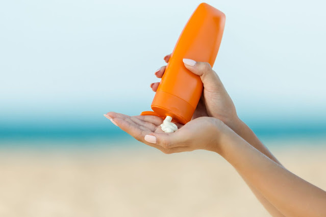 Beach background. Foreground: pair of hands holding an unlabled orange bottle of lotion, presumably sunscreen.