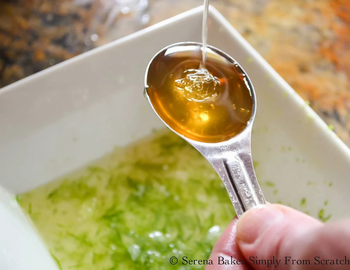 Lime and lime zest in a bowl with honey being poured into a measuring spoon over the top.