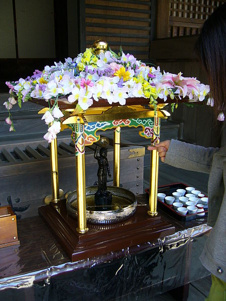 a small palanquin sitting in a temple hall is covered with flowers; inside stands a small black statue of a figure raising the finger of one hand up, and the finger of the other hand down