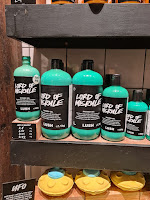A photo of a light brown rectangular shelf filled with a row of cylindrical bottles filled with green liquid with a black lid all with black rectangular labels that say Lord of Misrule shower gel in white font on a bright background