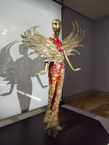 Photo of a red and gold brocaded woman's outfit on a gold-colored mannequin, with gold wing-like appendages attached to her front, one going to the left, one to the right. The lighting casts a dramatic shadow on the wall behind the mannequin.
