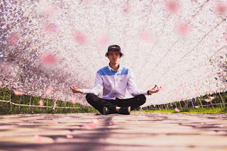 A man sits cross-legged, arms out in a stereotypical "meditation" post, on a stone path under a hemicylindrical trellis over which pink flowers or leaves have been trained.