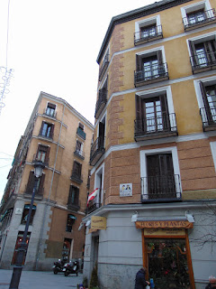 Lla placa de cerámica con el nombre de la calle en la esquina de un típico edificio con planta baja de granito y plantas superiores de color ocre con balcones enmarcados en blanco.