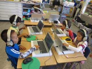 Students sit at computers doing work with headphones on