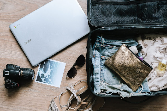 The image shows a travel flatlay. In the image, there are polaroid photos, sandals, a camera and a laptop. They are sitting on a wooden desktop next to an open suitcase filled with clothing