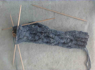 A sock-in-progress in worsted-weight yarn on double-pointed needles.  A cable pattern runs down the front leg and instep of the sock.  A stitch marker with a silver rose charm is clipped in at the back of the sock.