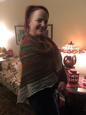 A pretty girl with a magenta-dyed undercut, standing in a sparsely decorated living room, with a big squishy colorful shawl draped over her shoulders. The off-white lace edge is a little crumpled, but the zigzag pattern still shows okay.