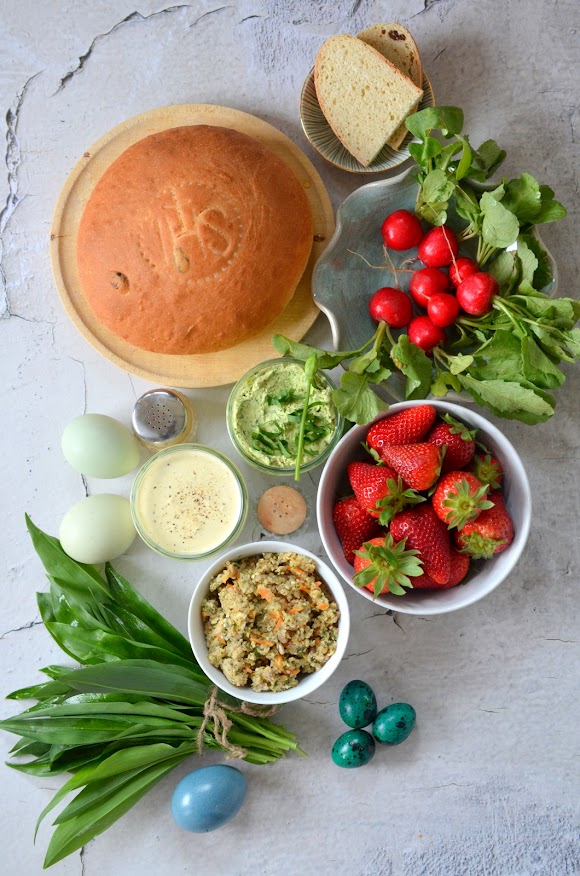 Die Zutaten für einen bunten Veggie-Weihkorb zum Osterfest liegen auf einem grauen Untergrund: Osterbrot, Radieschen, frische Aufstriche, gefärbte Eier, Bärlauch und Erdbeeren.