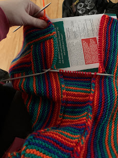 A strip of striped garter stitch, bordered by elongated Vees, being worked between two perpendicularly arranged pieces of striped garter stitch. All of the stripes are in the same red, bright orange, green, and royal blue.