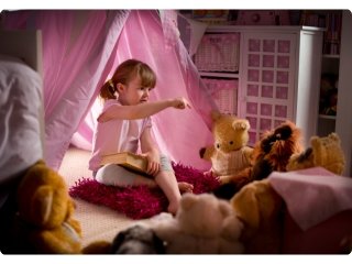A girl sits at the entrance of her room tent, having a conversation with her soft toy audience who are seated in a circle in front of her.