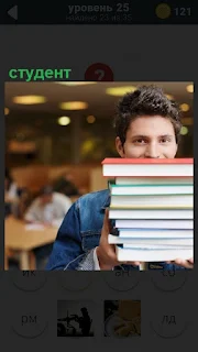 Парень студент в библиотеке несет в руках высокую кипу книг сдавать