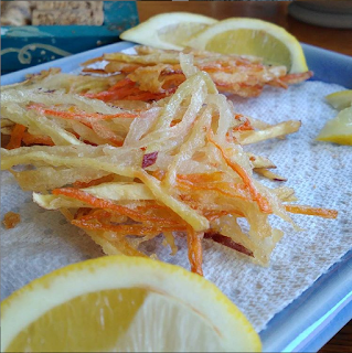 Julienned root vegetables lightly battered and then deep friend in small bunches. The end product sits on a paper towel with lemon wedges. 