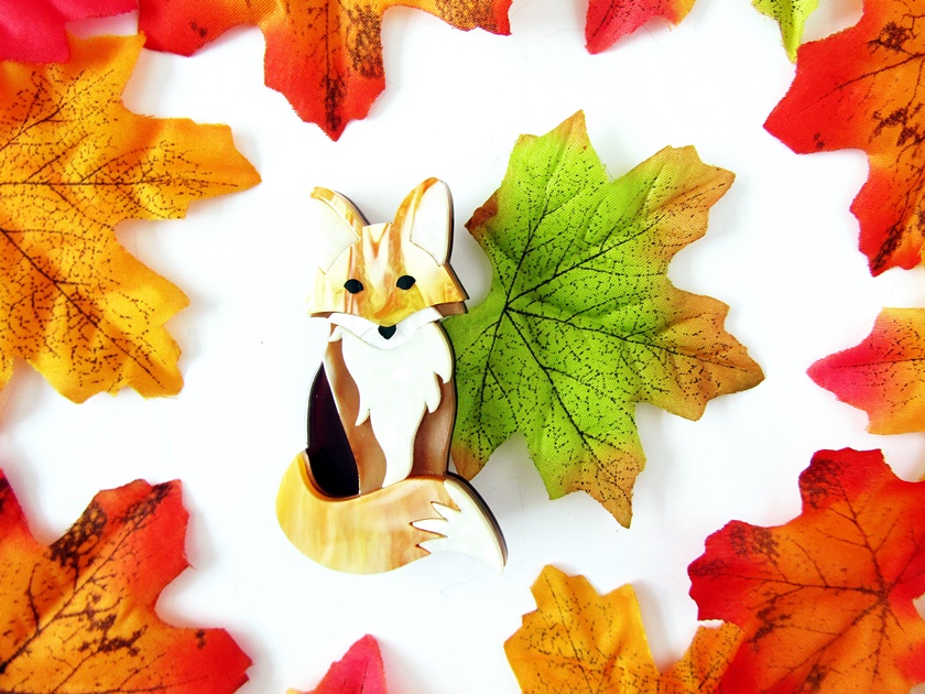 A photo of Erstwilder's 'Mini Mulder' red fox brooch taken from above. The photo shows a golden-brown and white sitting resin fox brooch facing forwards. It sits on a white background surrounded by fiery faux autumn leaves