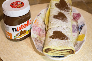 Foto di rotolo con la nutella con cuori di cacao per la festa di san valentino. il rotolo con la nutella, anche chiamato girella, è un dolce semplice da preparare e di sicuro gradimento. è ottimo per la prima colazione o per la merenda del pomeriggio, ma può essere gustato anche come dessert alla fine del pasto. nella foto, adagiato su vassoio e su superficie rustica, con barattolo di nutella in sfondo. 