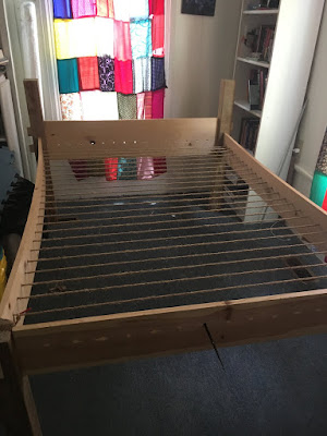 A wooden bed fram with 4 by 4 posts and wide, thin rails, head, and footboard with heavy jute rope laced so tightly from side to side that the side rails are bowed in. Sunlight pours through the patchwork curtain over the window at the head of the bed.