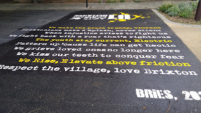A poem in yellow and white on black asphalt: "we make the place trendy, vibrant Brixtonians make a Splash, never silent. When injustice arises to fight us we fight back with a roar that's righteous. The youth stay current, Electric Pattern up 'cause life can get hectic. We grieve loved ones no longer here. We kiss our teeth to conquer fear. We rise, Elevate above friction. Respect the village, love Brixton. BRiES.
