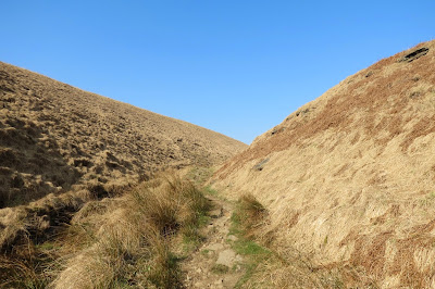 Pale moorland grasses on the side of the clough and a narrow path leading into this small valley. Cloudless blue skies above.