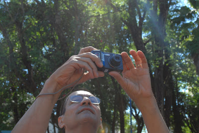 A imagem mostra os braços de Paulo acima de sua cabeça, segurando a câmera e olhando em direção a ela. Ao fundo aparecem as copas de algumas árvores e reflexos do sol.