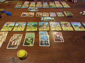 A game of Majesty: For the Realm in progress. The photo is taken from the point of view of one player, looking at his tableau. The location cards are laid out in front of him, with three millers, two brewers, two witches, two guards, and one knight under their respective location cards. A stack of yellow coin tokens sits nearby. Above the location cards, the worker card holds five meeples. The row of character cards is above that. In the background, the other player's tableau can be seen. A variety of coin tokens in various colours can be seen off to one side. A pile of white meeples can be seen on the other side.
