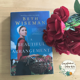 a wooden table with the book a beautiful arrangement by Beth Wiseman next to a bunch of red roses