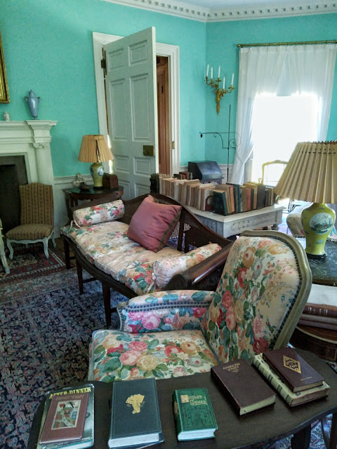 Another View of the Drawing Room, including a floral couch and chair set, more books, more chairs, more side tables, more lamps, more vases, and the corner of the fireplace.
