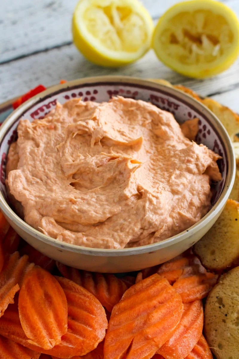 Side view of Smoked Salmon Dip in a tan bowl on a blue plate with bagel chips, carrot chips, and bell pepper pieces around it.