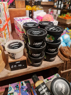 A bunch of cylindrical black pots stacked on top of each other with a circular black label that says lovely hand balm lush in white font on a large light brown shelf on a bright  bsckground