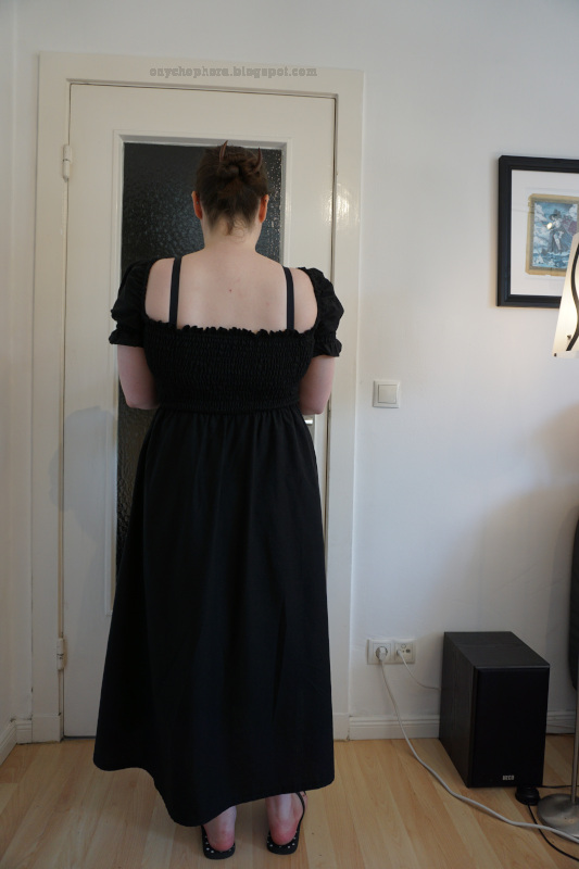 Eine weiße Frau steht in einem schwarzen, kurzärmeligen Kleid vor einer weißen Tür mit dem Rücken zur Kamera.