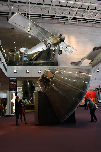 In a brightly lit space with maroon carpet sits the gumdrop shaped Apollo 11 Capsule, tan in color and looking old but majestic on its mount of black and gold.  Little Star (a smiling yellow star) sits on top of the mount and overhead "The Spirit of Saint Louis" can be seen.  The airplane is silver with small black tires, a propeller and the name of the aircraft on the side of the body near the nose.