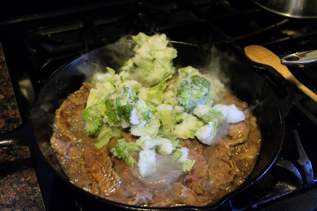 The frozen broccoli being added to the skillet with the beef.