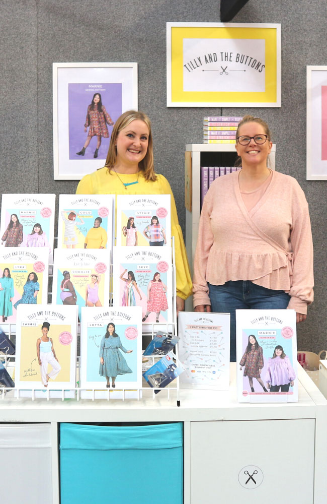 Two Team Buttons employees stand smiling at the Tilly and the Buttons stand, surrounded by sewing patterns and sewing books