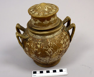 A pale brown pot with a lid with delicate white patterns on it, including flowers