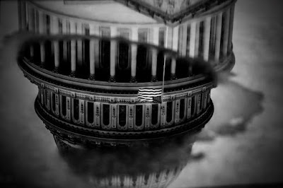 black and white photograph of the US Capitol and American flag reflected in a puddle of water on the ground, Washington, DC, January 3, 2021