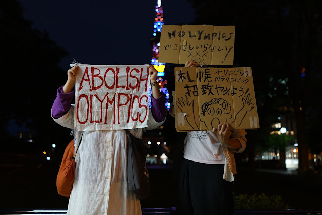 어두운 밤 조명으로 장식된 구조물 앞에서 두 사람이 천과 골판지로 만든 피켓을 들고 서 있다. 피켓에는 영어와 일본어로 '올림픽을 폐지하라', '올림픽은 어디에도 필요없다', '삿포로 올림픽 유치를 멈춰라' 등의 내용이 적혀있다.