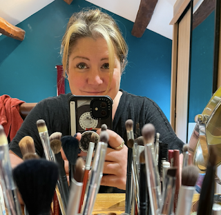 A photo of me - highlighted brown hair, pulled back into a knot, sitting in front of my makeup mirror. The photo is of the reflection. I'm neither smiling or scowling. Just looking at the mirror and wearing a black top.