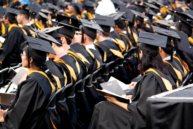 A large group of graduates, seen from behind and to the side, seated in rows of chairs at a commencement ceremony. They are all wearing black graduation gowns and mortarboard caps with gold stoles. Several graduates are looking towards the front, and some are holding programs or books.