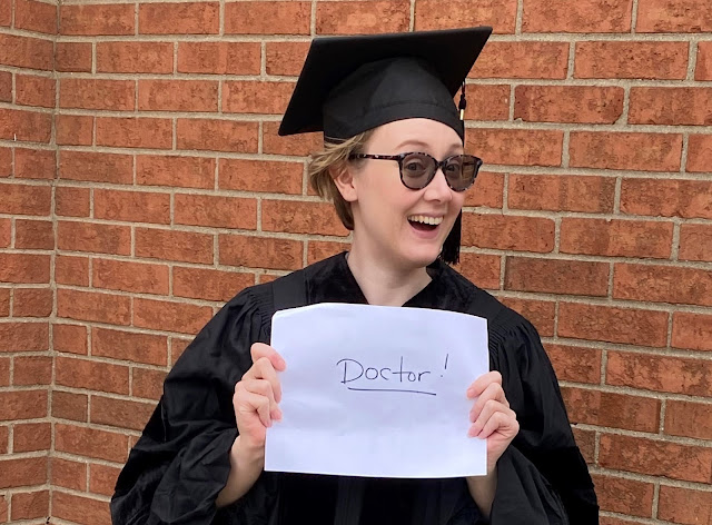 Me wearing a graduation cap and gown and holding a piece of paper that says "Doctor!"