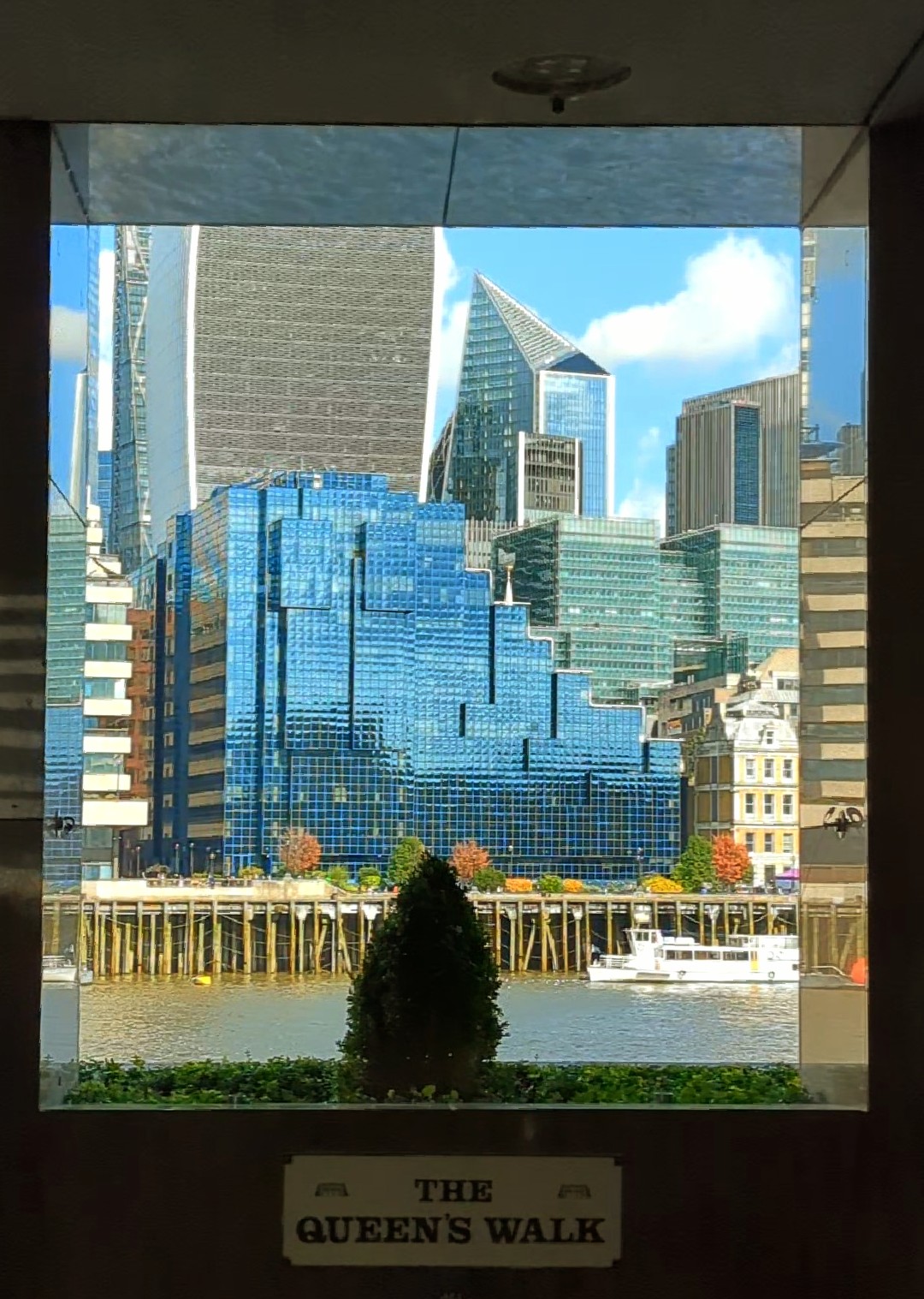 A large square opening in the dark looking across the River Thames to the many shapes of tall glass covered buildings on the other side. They are reflecting the blue / green of the sky. A small conical tree sits in the openings window box, is it real who knows. A sign underneath reads, "The Queen's Walk"