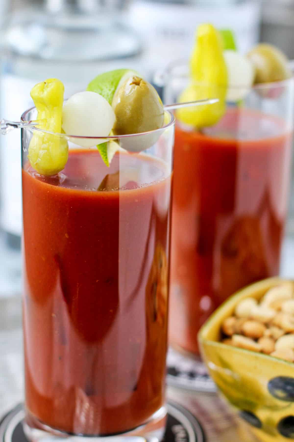 Bloody Marys in two glasses with peanuts in a bowl on the side.