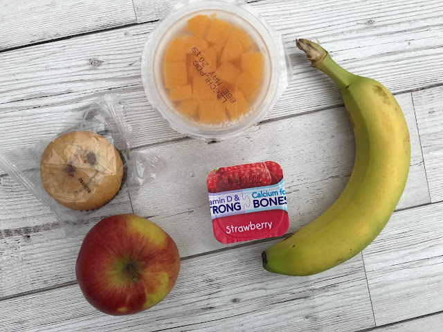 A photo of food laid out, banana, apple, muffin, fruit pot, yogurt