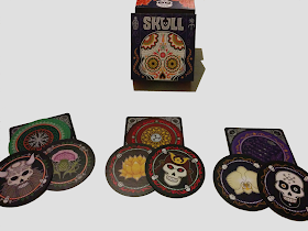 The box, decorated with a colourful stylized skull, with three sets of cards. Each set (one green, one red, and one purple) displays two cards face down on the score mat and two cards face up. The score mat is square, and is as wide as the cards, which are round. The back of each card has an intricate pattern. The two face up cards in each set show one flower and one skull. The green cards flower is a thistle, the red cards have a lotus, and the purple cards have an orchid. The green cards' skull is horned like a stereotypical Viking, the red cards' skull is wearing a samurai helmet, and the purple skull has a geometric design on the forehead and the jawbone painted black.