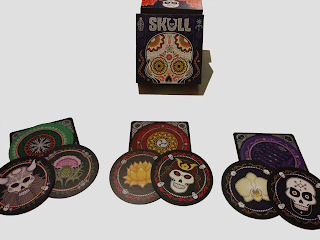 The box, decorated with a colourful stylized skull, with three sets of cards. Each set (one green, one red, and one purple) displays two cards face down on the score mat and two cards face up. The score mat is square, and is as wide as the cards, which are round. The back of each card has an intricate pattern. The two face up cards in each set show one flower and one skull. The green cards flower is a thistle, the red cards have a lotus, and the purple cards have an orchid. The green cards' skull is horned like a stereotypical Viking, the red cards' skull is wearing a samurai helmet, and the purple skull has a geometric design on the forehead and the jawbone painted black.