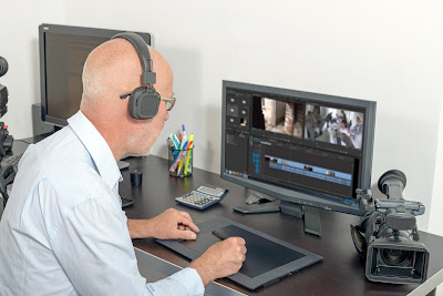 Pic of man sitting in front of video editing programme on screen and handheld camera on desk