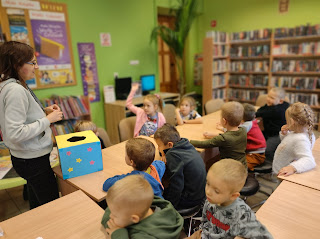 Dzieci siedzą przy stolikach, jedna z dziewczynek zgłasza się do odpowiedzi. Obok stoi pani bibliotekarka przed którą leży kolorowe kartonowe pudełko.